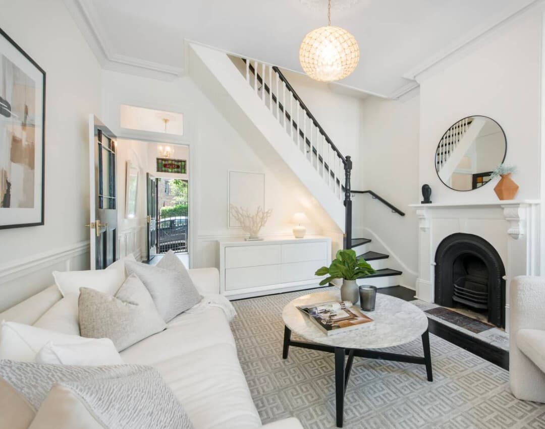 Bright, modern living room featuring a white sofa, marble coffee table, fireplace, and staircase.