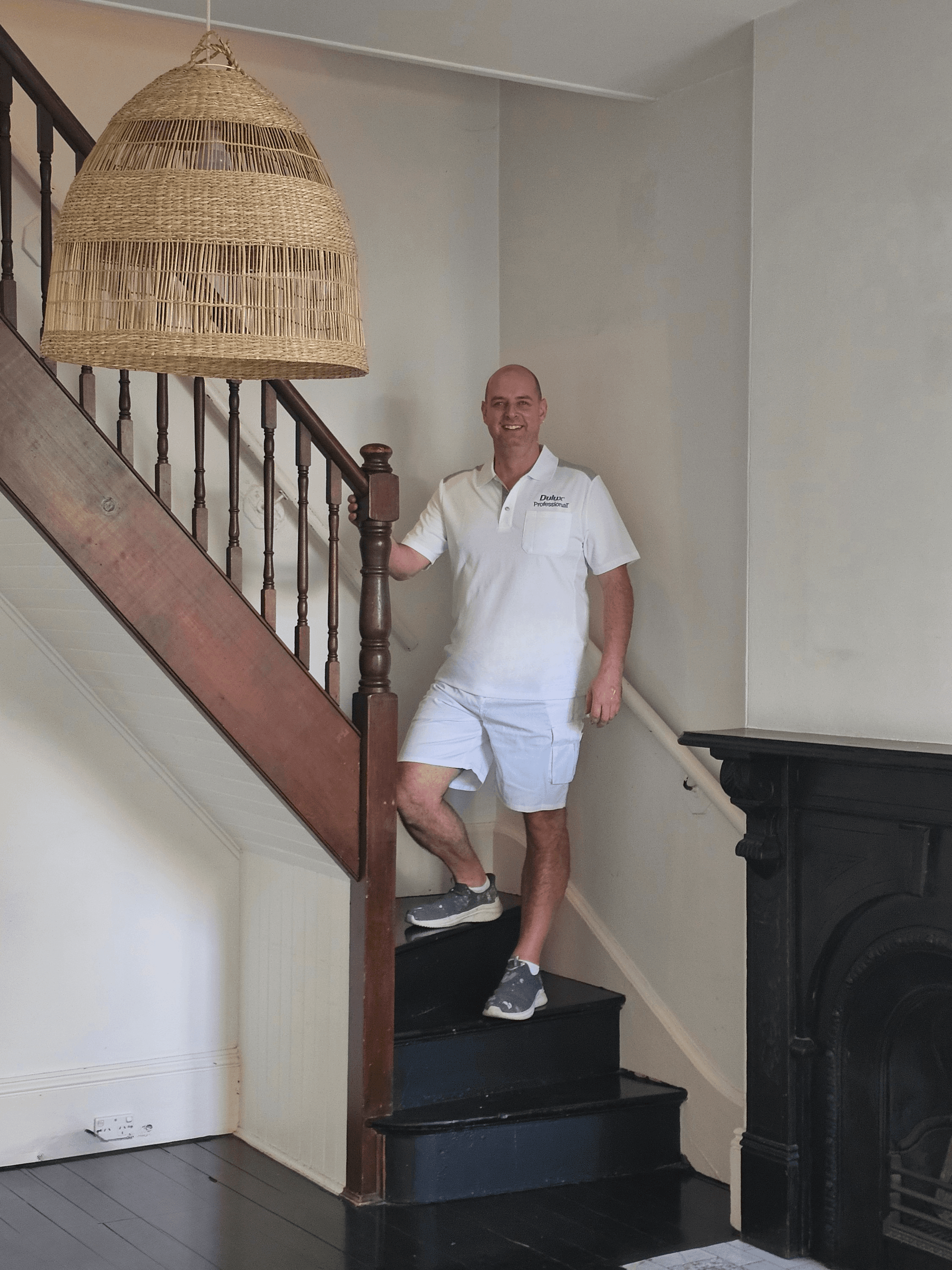 Smiling man in white Dulux uniform stands on stairs under a large woven wicker lamp.