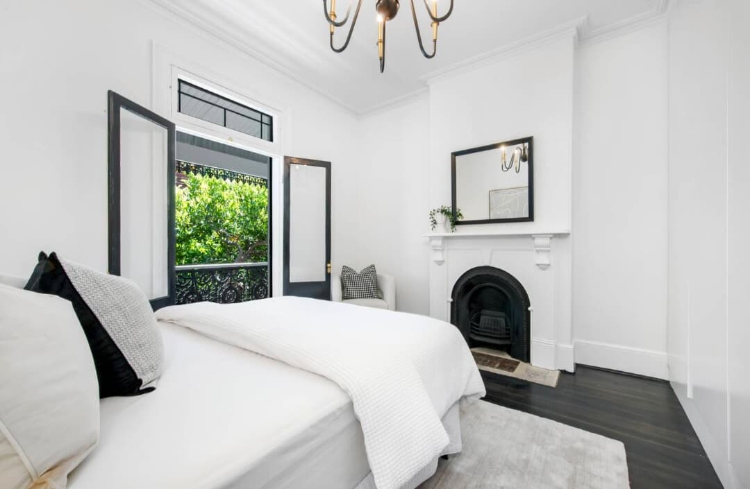 Bright white bedroom with a fireplace, mirror, and open balcony doors overlooking green trees.